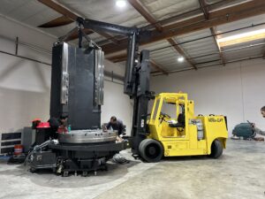 Yellow forklift lifting large industrial machine indoors.