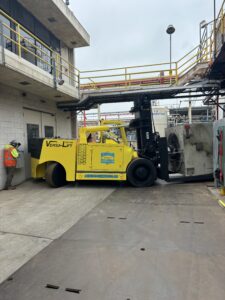 Yellow forklift moving large equipment in facility.